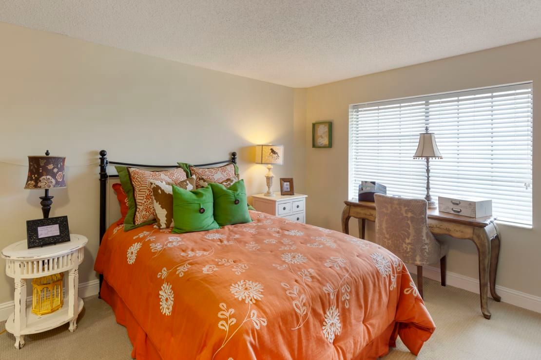 A cozy bedroom with an orange bedspread and green pillows, featuring a lamp on a nightstand and a desk by the window.