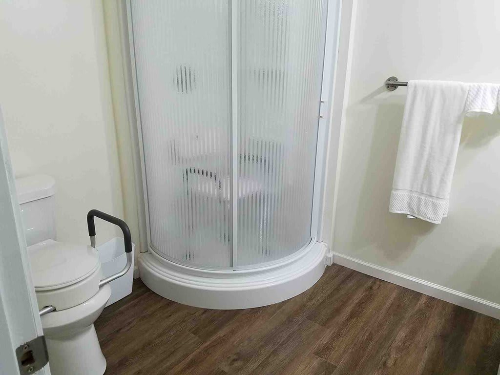 A bathroom featuring a corner shower with frosted glass, a toilet with a handrail, and a towel rack on a wooden floor.