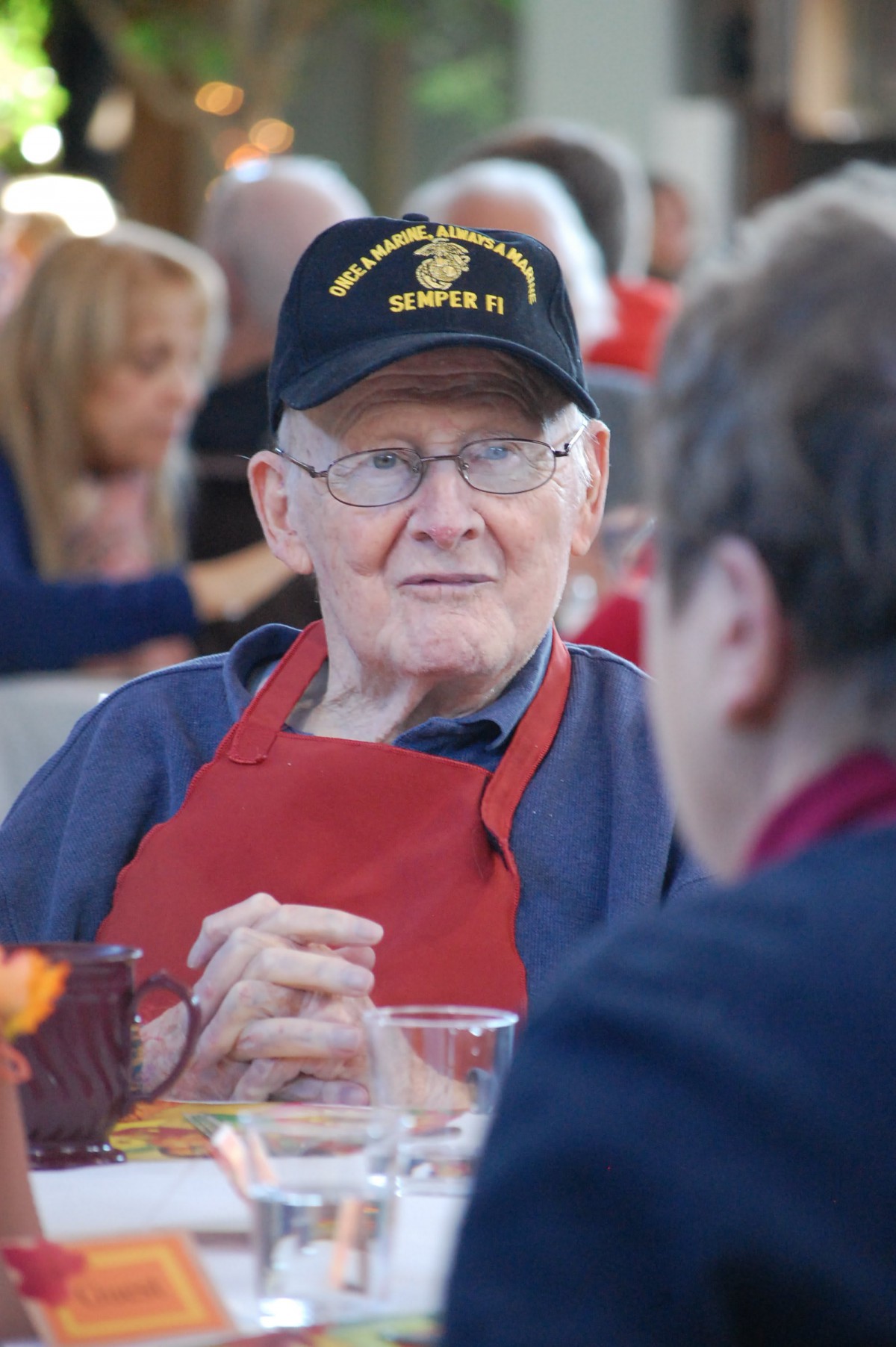 An elderly man wearing a hat and red apron engages in conversation at a social gathering.