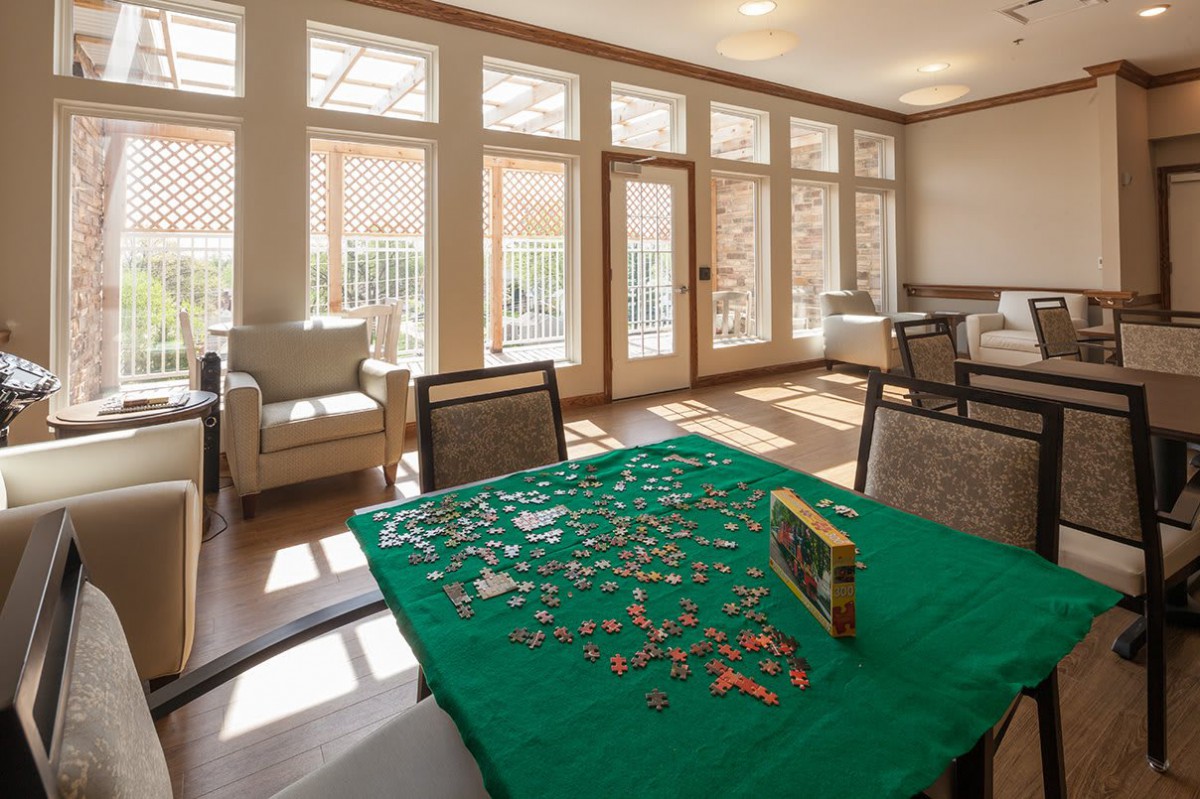A sunlit room with a puzzle in progress on a table surrounded by chairs.