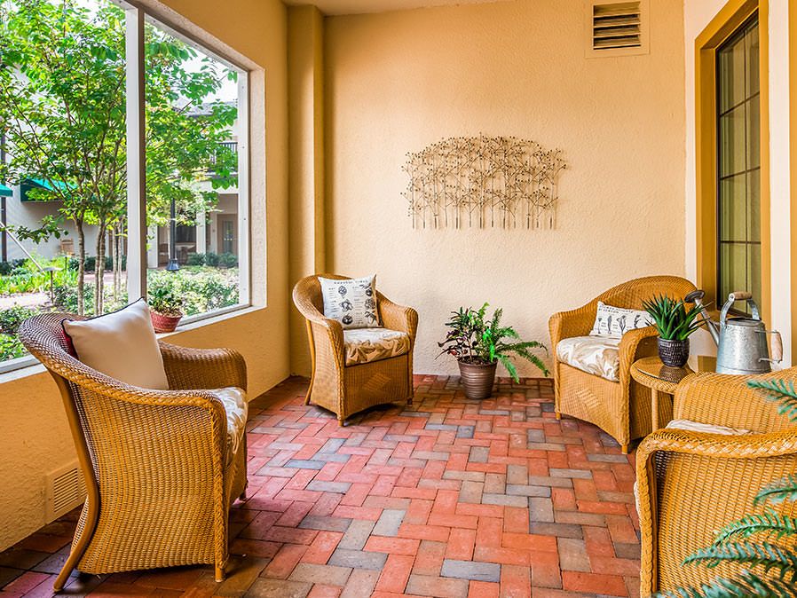A cozy sunroom with wicker chairs, indoor plants, and brick flooring, bathed in natural light.