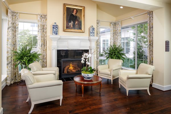 A cozy living room with a fireplace, surrounded by armchairs and a coffee table, decorated with plants and a portrait above the mantel.
