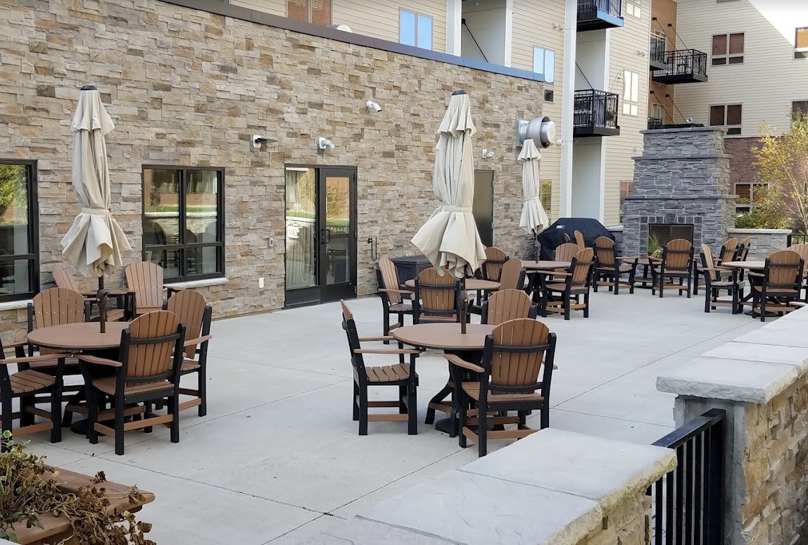 An outdoor patio area with tables and chairs, set against a brick wall, featuring closed umbrellas.