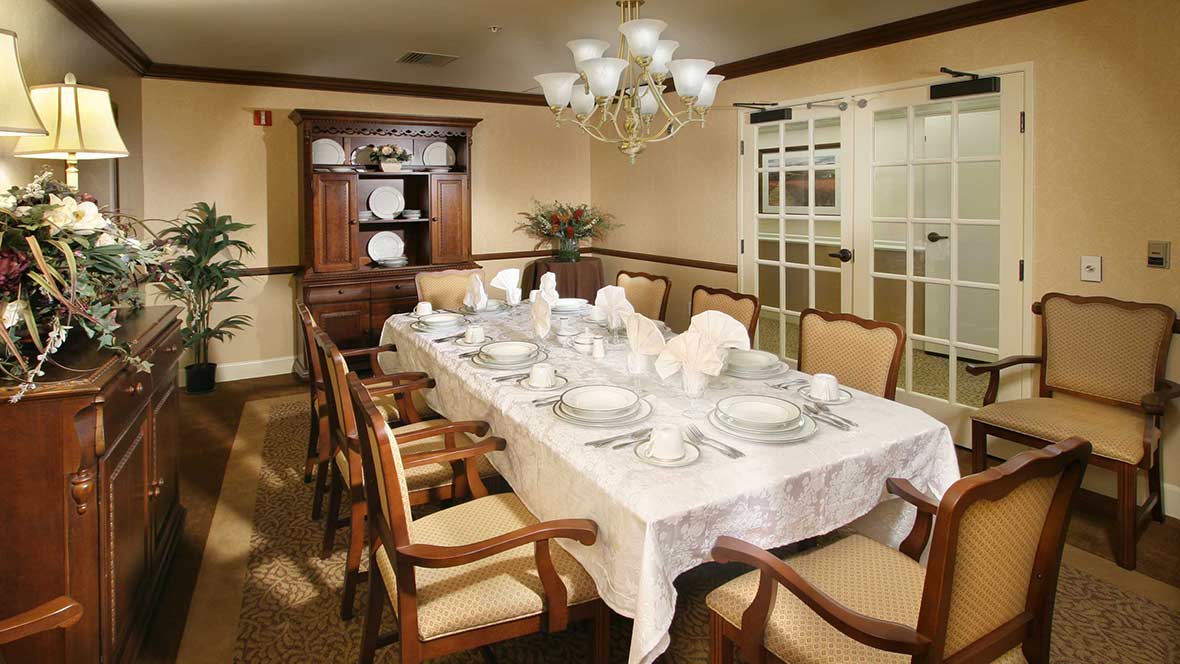 An elegantly set dining room with a long table, surrounded by chairs and adorned with fine tableware under a chandelier.