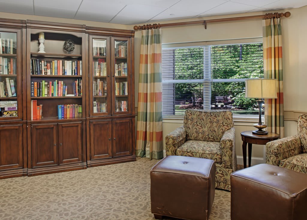 A cozy living room with a bookshelf, armchair, and ottomans near a window.