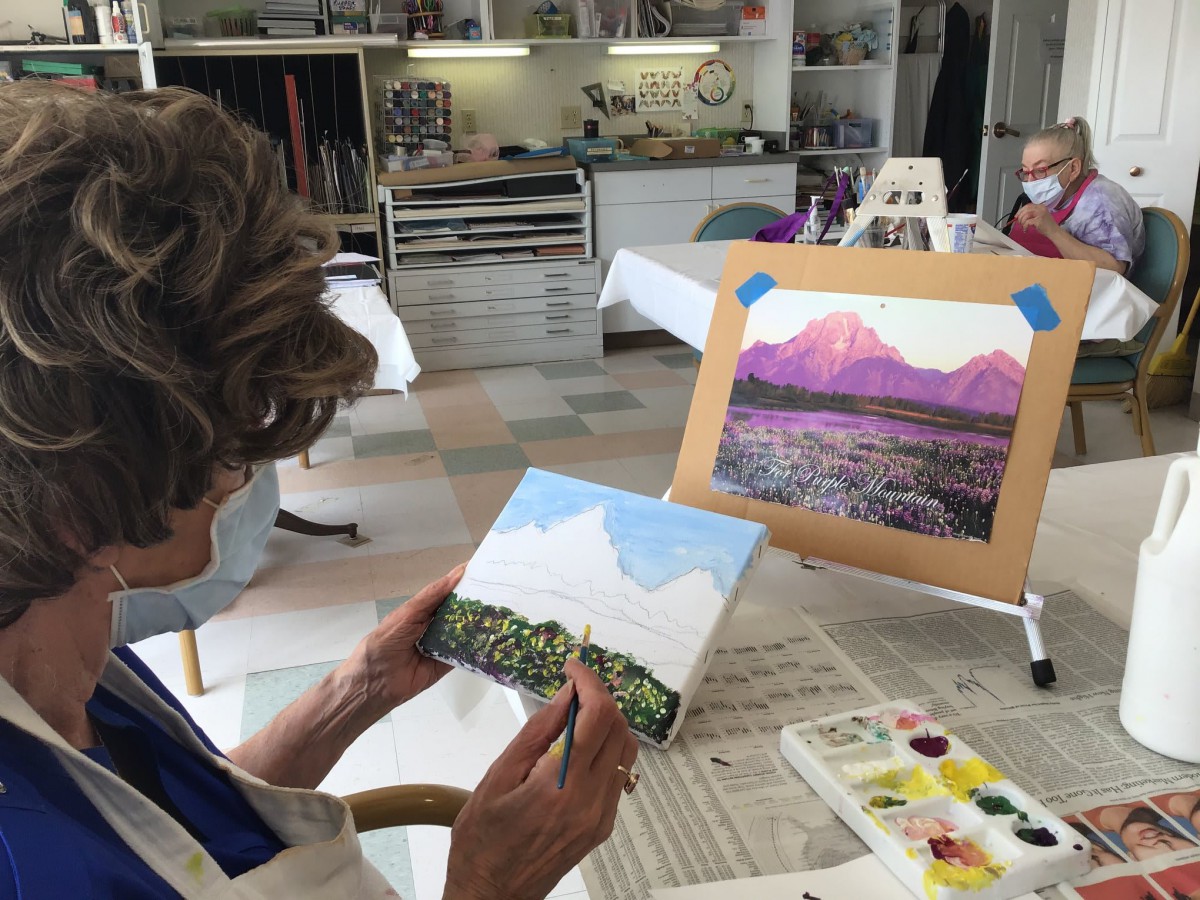 An artist working on a landscape painting in an art studio, with a completed mountain scene on display.