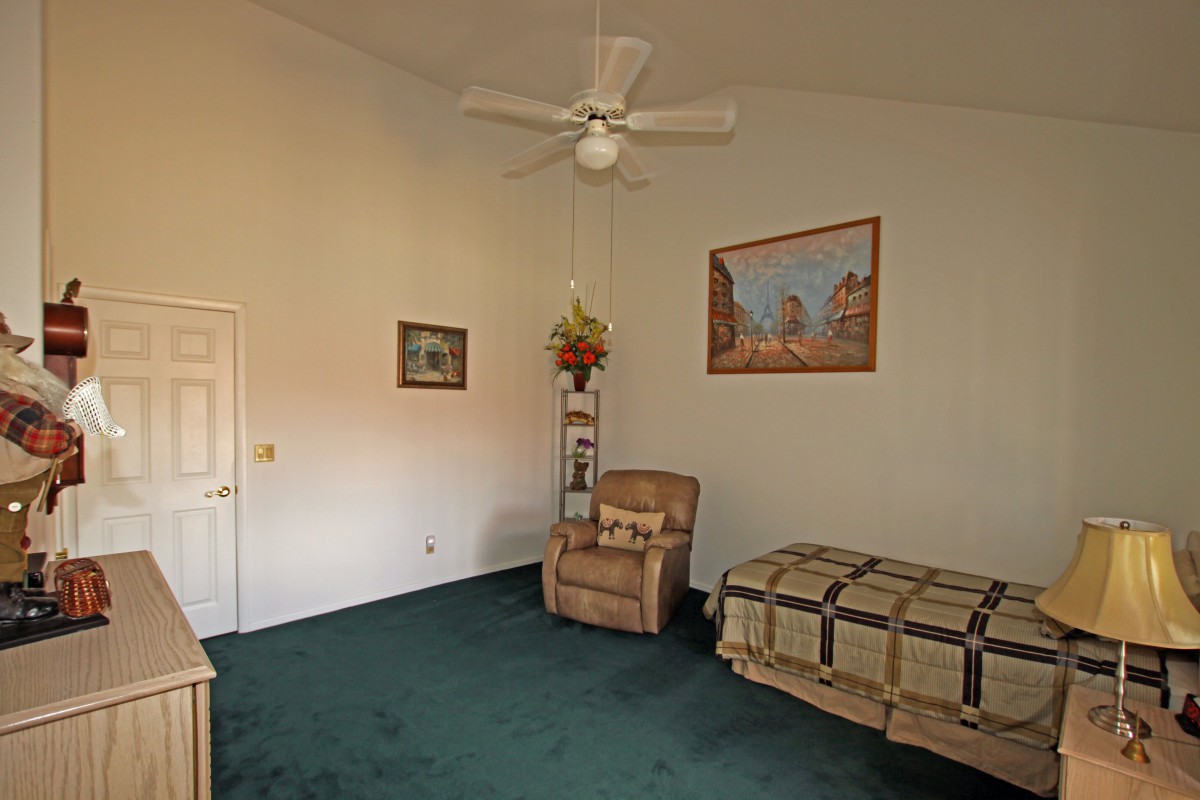 A simple bedroom with a single bed, armchair, ceiling fan, and decorative wall art.