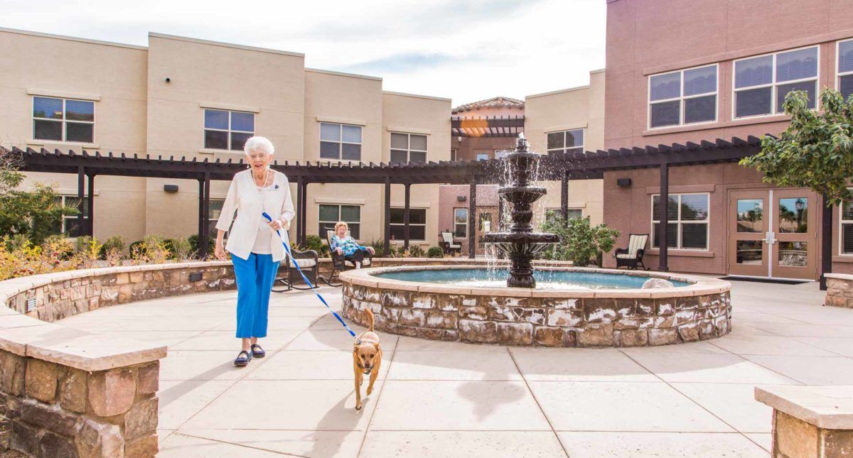A senior woman walks her dog in a courtyard with a fountain at a retirement home.