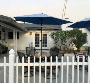 A cozy backyard patio area with tables, chairs, blue umbrellas, and a white picket fence.