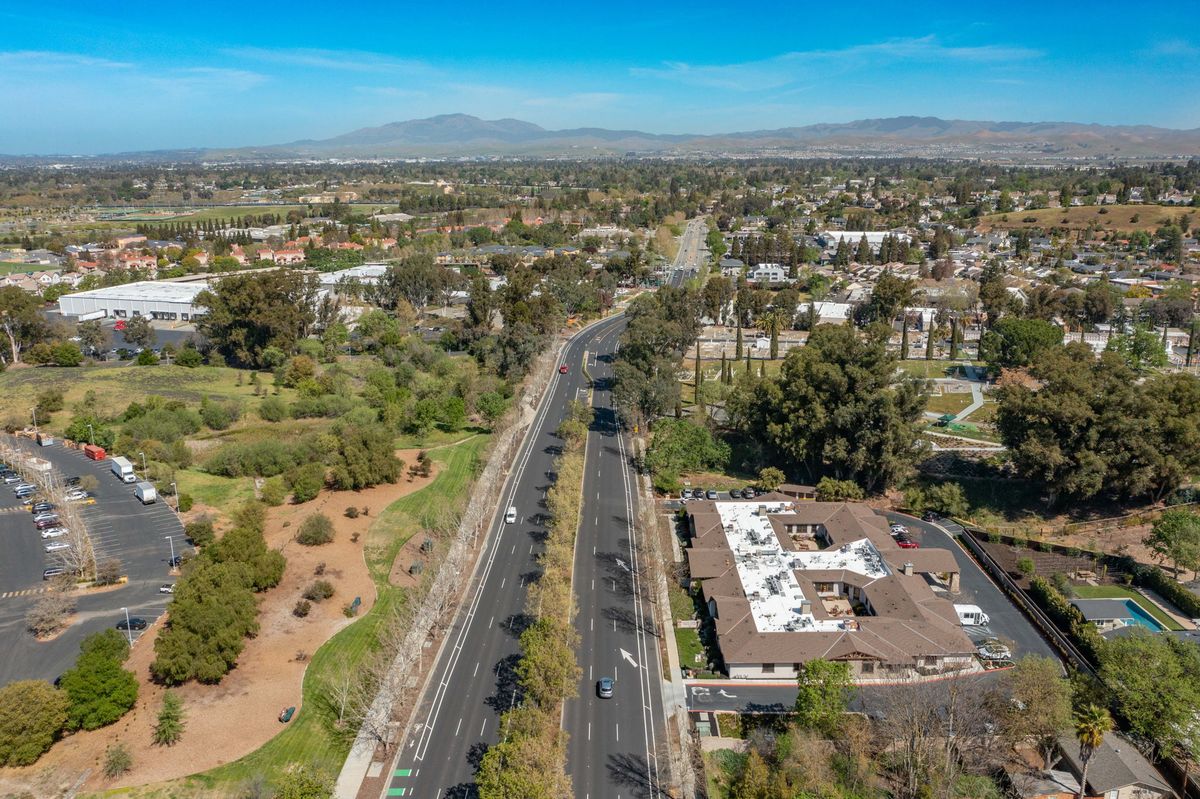 Aerial view of an urban landscape featuring a road lined with trees, buildings, and mountains in the background under a clear blue sky.