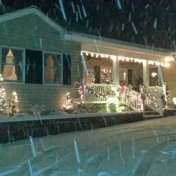 A cozy house adorned with Christmas lights, gently blanketed by falling snow on a winter night.
