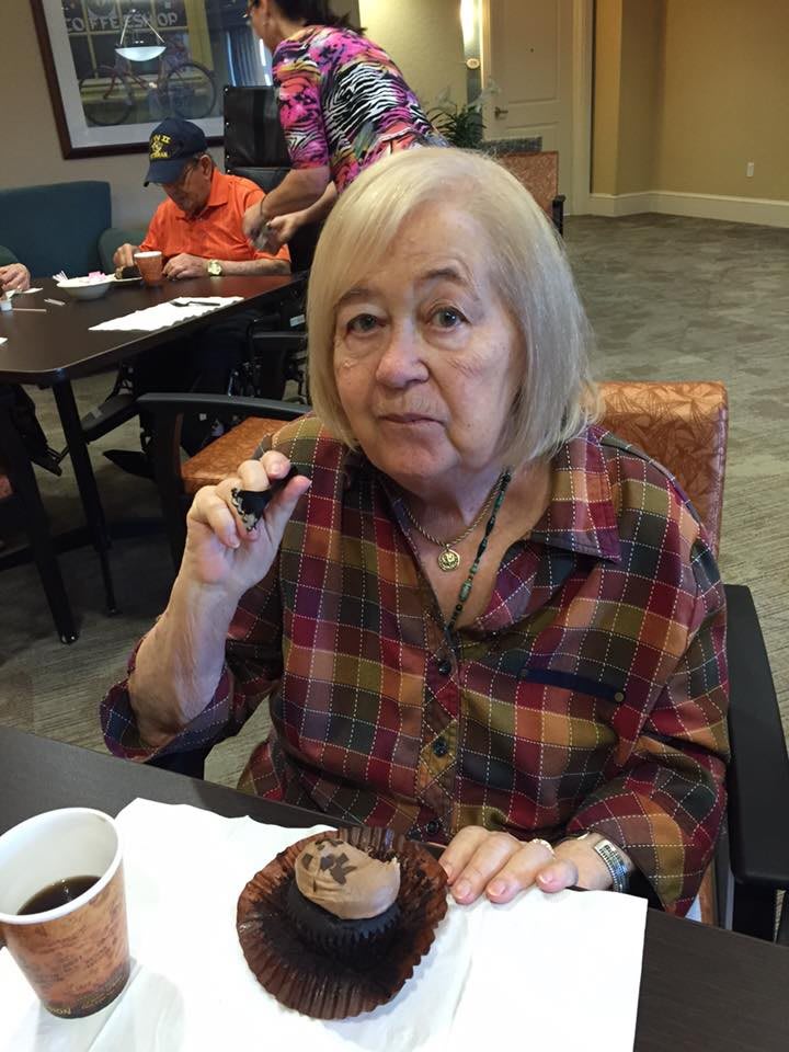 An elderly woman enjoying a cupcake while sitting at a table indoors.
