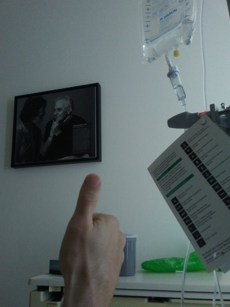 A person gives a thumbs-up in a hospital setting with an IV drip and medical chart visible on the side.