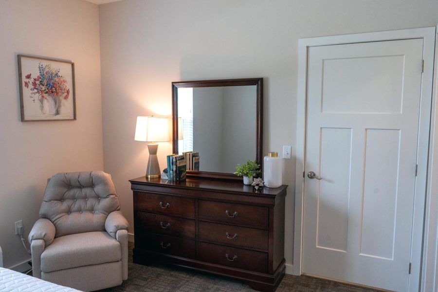 A cozy bedroom corner featuring a dresser with a mirror, a lamp, and an upholstered chair.