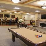 A recreation room with a pool table and people gathered at tables in a well-lit, cozy environment.
