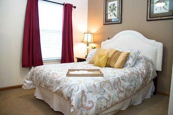 A cozy and elegantly decorated bedroom featuring a bed with patterned bedding, accent pillows, a bedside lamp, and burgundy curtains framing a window.