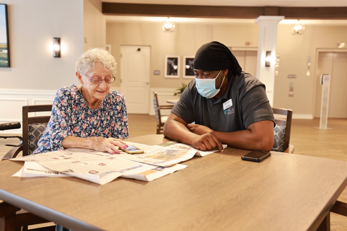 A caregiver and an elderly woman reading a newspaper together indoors.