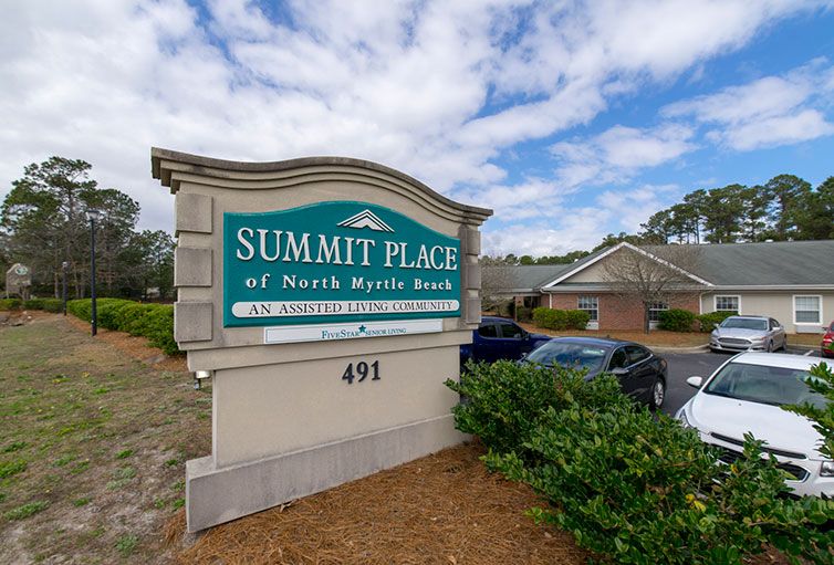 A sign for Summit Place of North Myrtle Beach, with a building and cars in the background under a partly cloudy sky.
