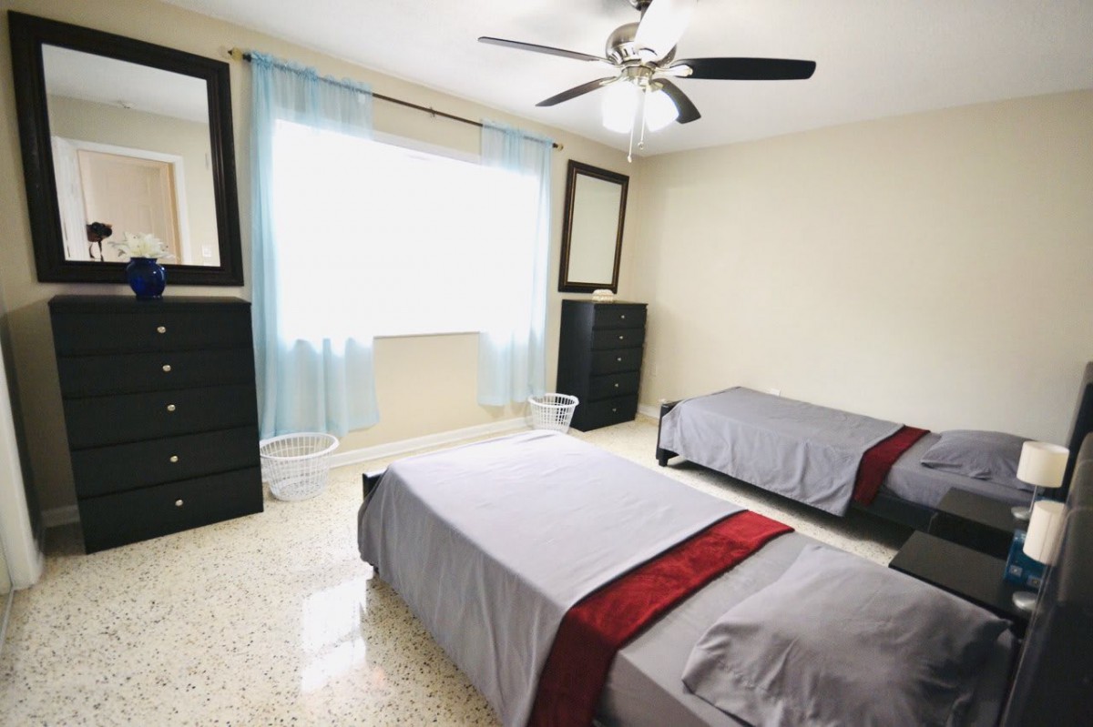 A neatly arranged bedroom featuring two twin beds with gray and red bedding, ample natural light from the window, and a ceiling fan for ventilation.