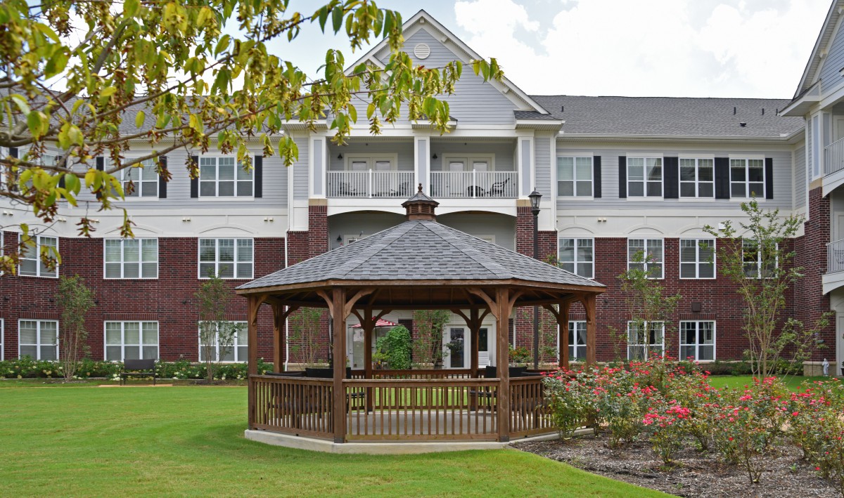 A wooden gazebo in front of a multi-story residential building with a well-maintained garden.
