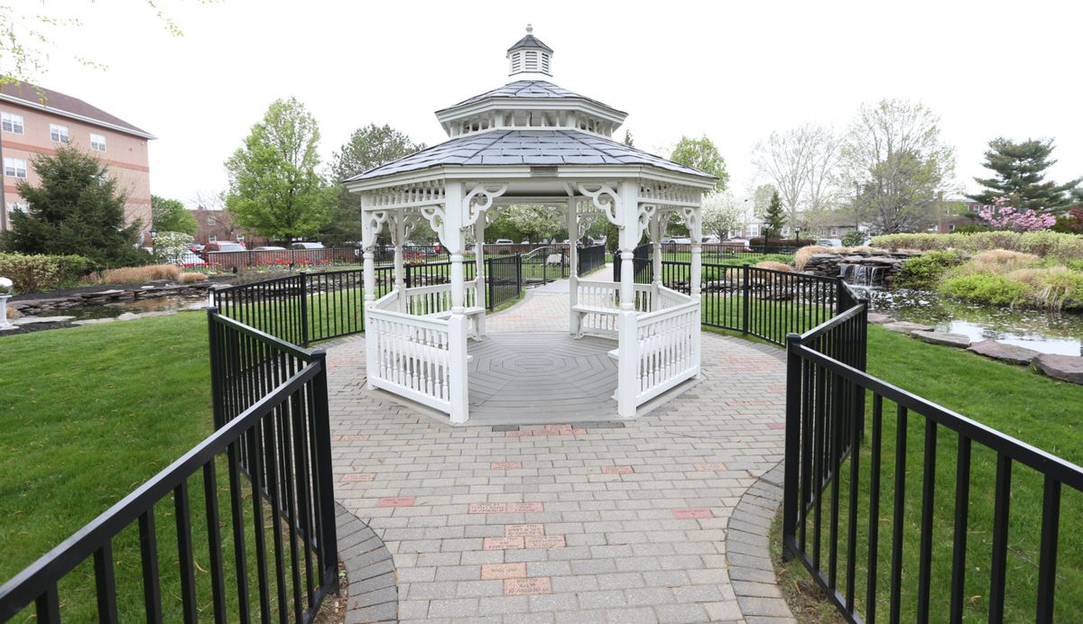 A white gazebo situated on a brick pathway in a park setting, surrounded by greenery.