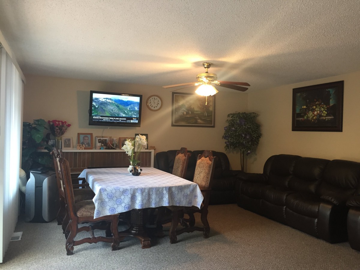 A cozy living room featuring a dining table, sofas, a television on the wall, and a ceiling fan.