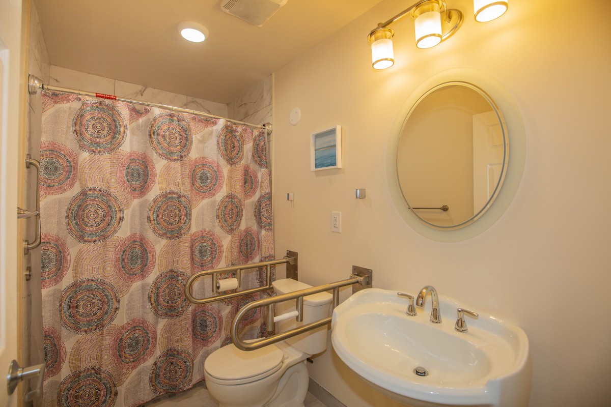 A well-lit bathroom featuring a sink, toilet with handrails, and a shower area.