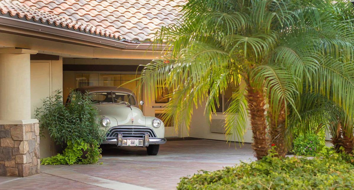 A vintage car parked in the driveway of a house surrounded by tropical greenery.