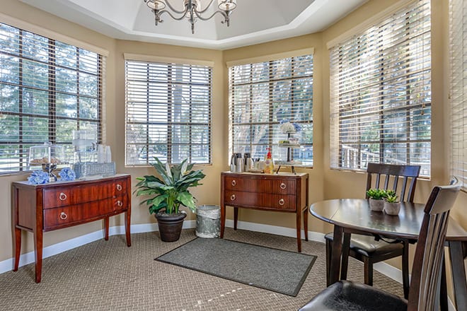 A cozy living room with large windows, wooden furniture, and a potted plant.