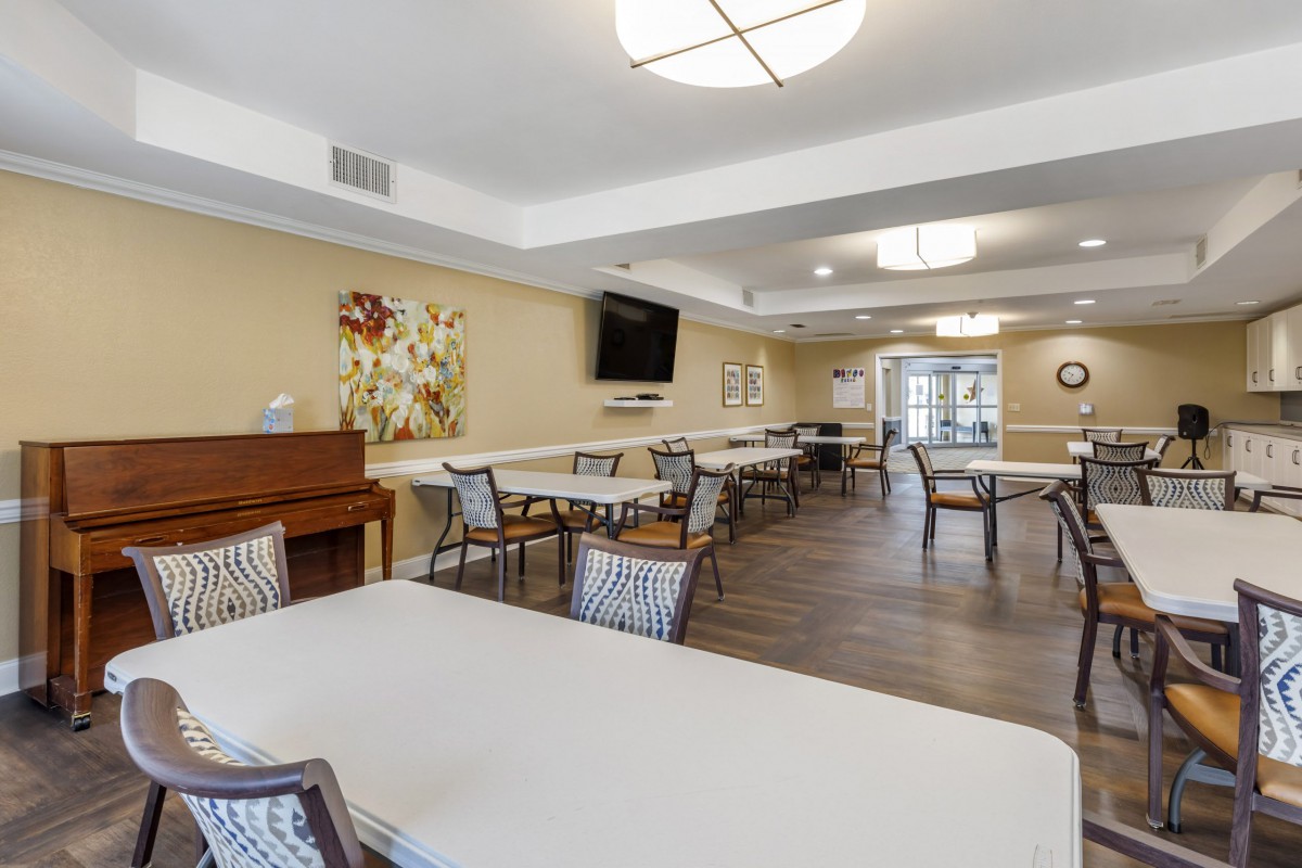A spacious dining area with multiple tables and chairs, featuring a piano and a mounted television on the wall.