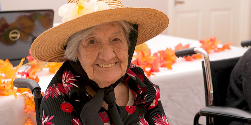 An elderly woman smiles warmly, wearing a straw hat and floral-patterned clothing.