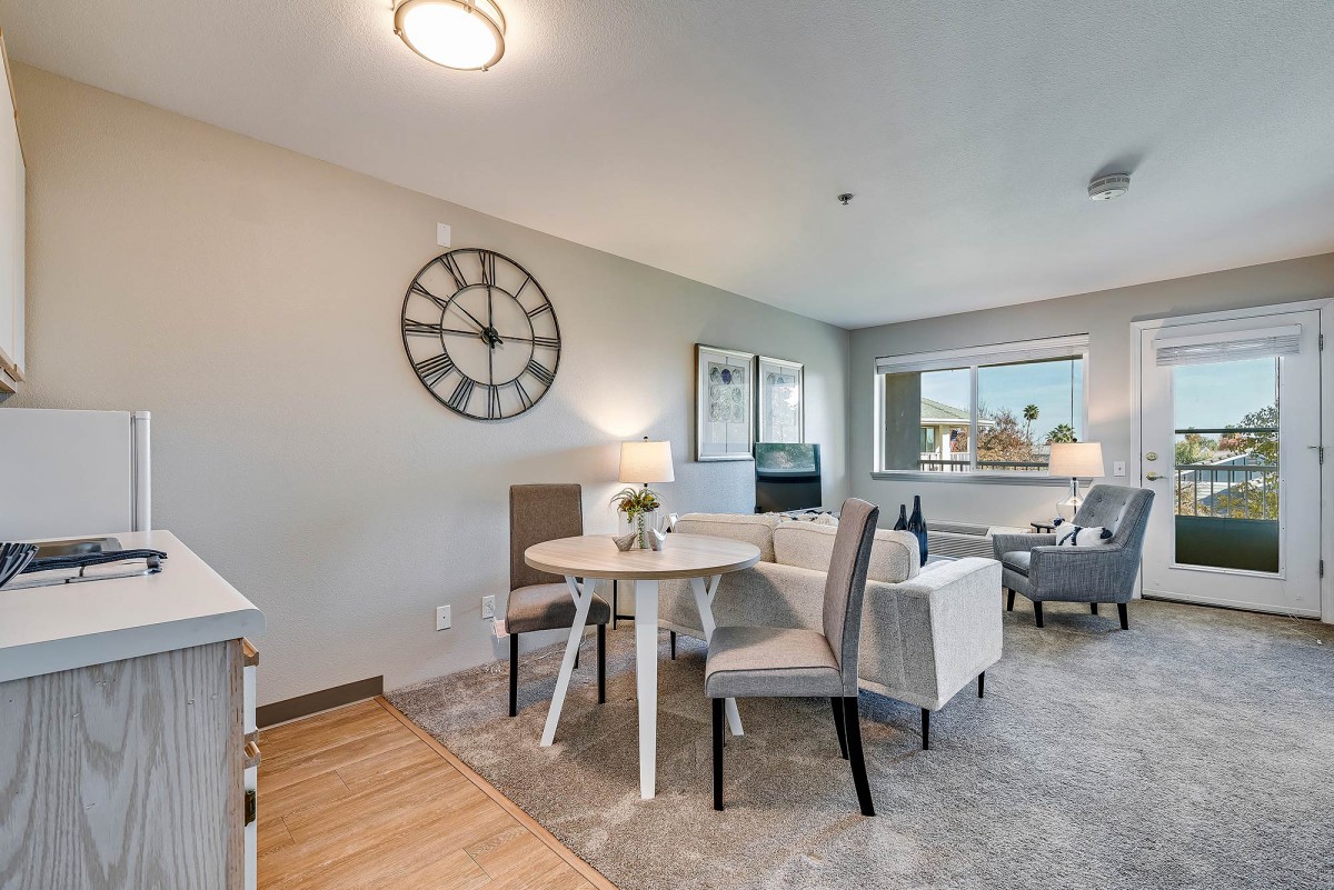 A modern living room and dining area with neutral tones, featuring a round table surrounded by chairs, a large wall clock, and ample natural light from the window.