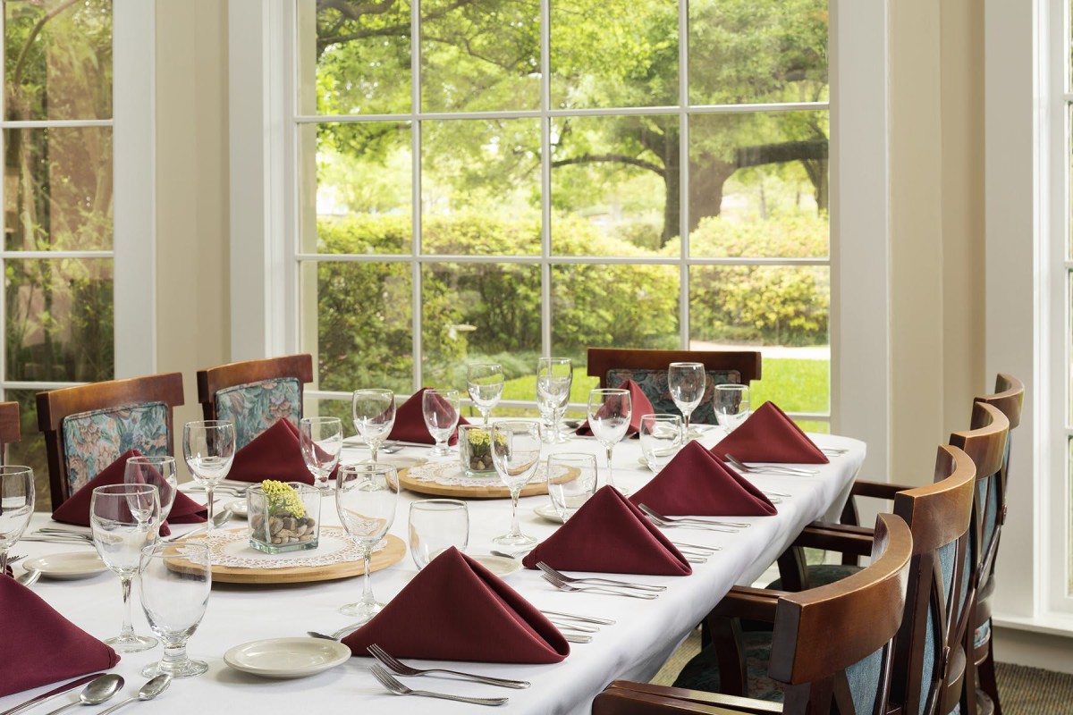 An elegantly set dining table with maroon napkins and wine glasses, bathed in natural light from a large window overlooking a garden.