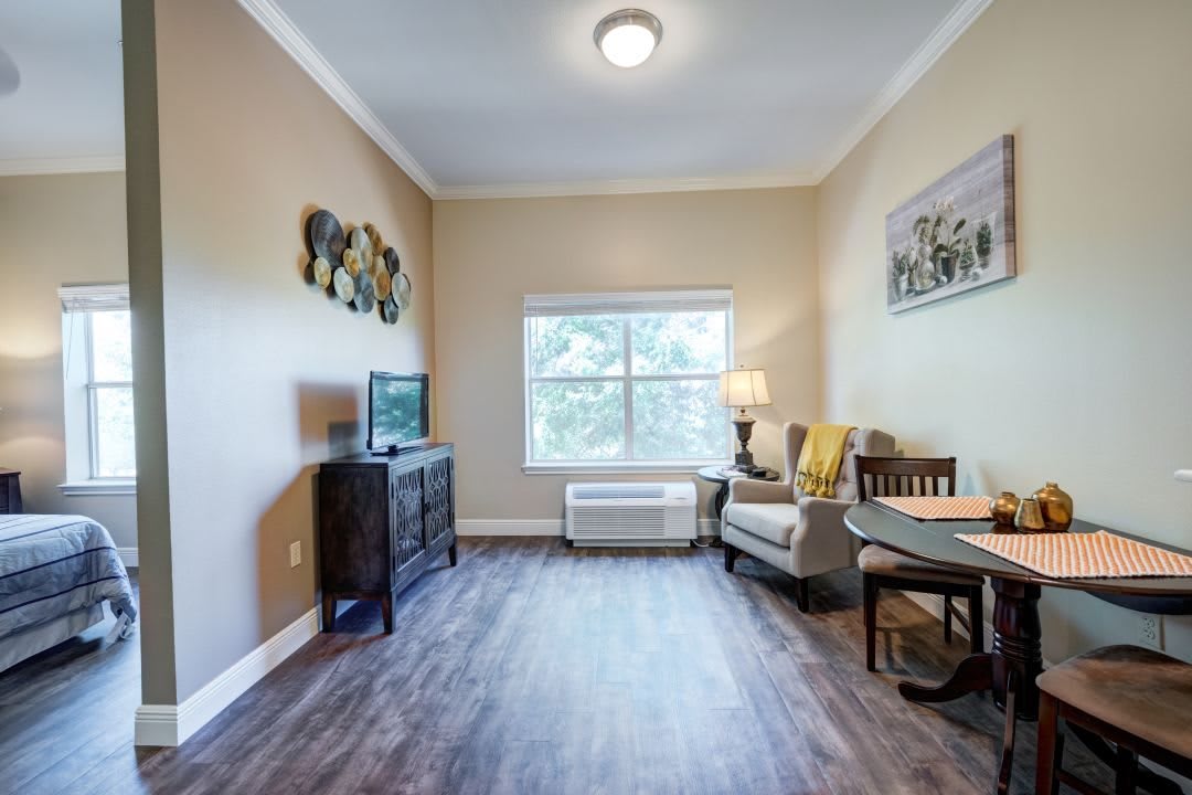 A cozy living room with a television, chair, and table, featuring a large window and decorative artwork.