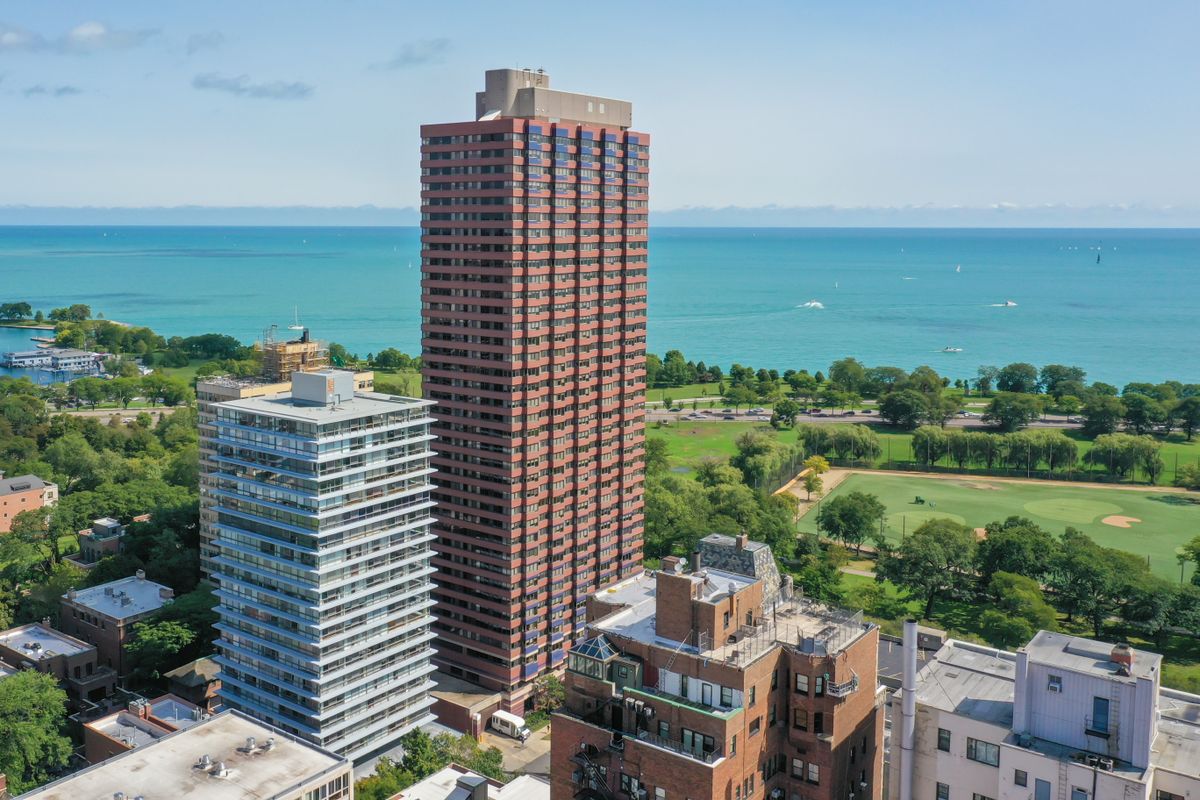 A large skyscraper stands tall near a lake and park, surrounded by smaller buildings in an urban setting.