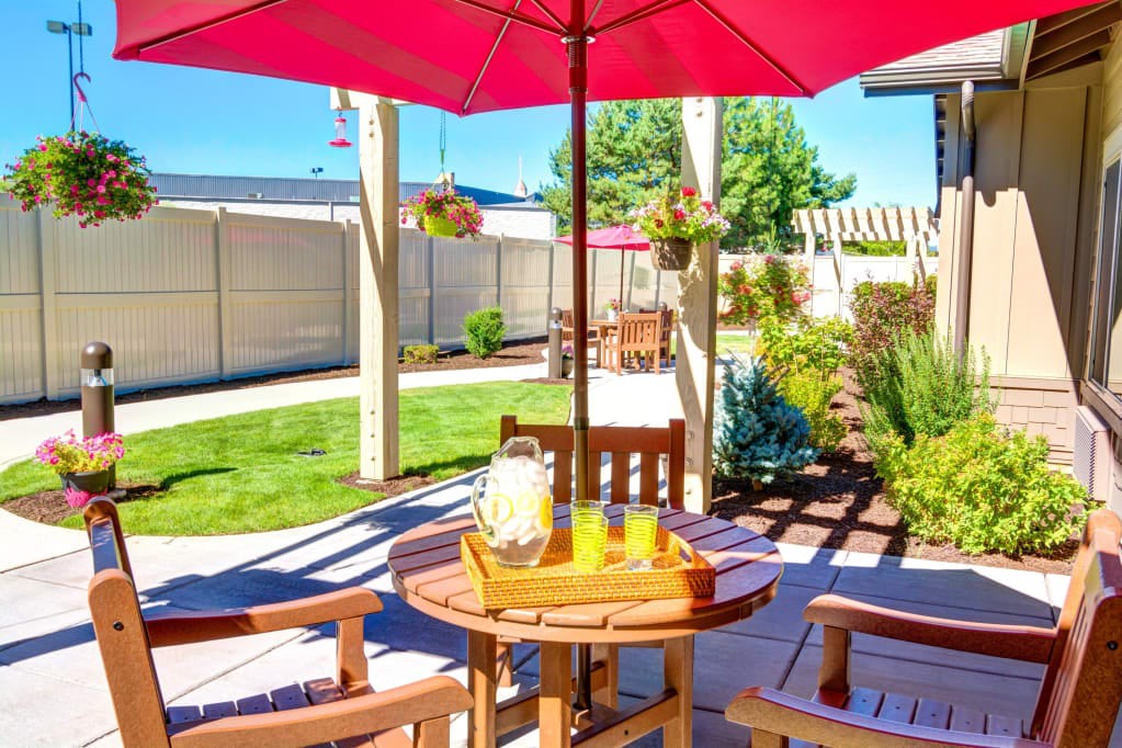 A sunny patio with a wooden table and chairs under a red umbrella, surrounded by a well-maintained garden.