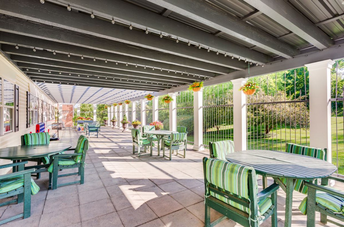A sunny outdoor patio with green furniture under a pergola, surrounded by hanging plants.