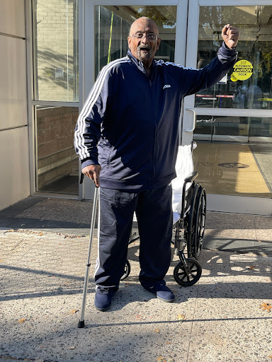 An elderly man stands outdoors, gesturing while holding a walking stick next to a wheelchair.