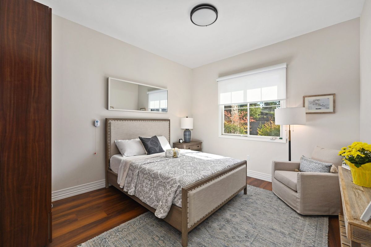 A modern bedroom with neutral tones, featuring a bed with pillows and a bedside table lamp.