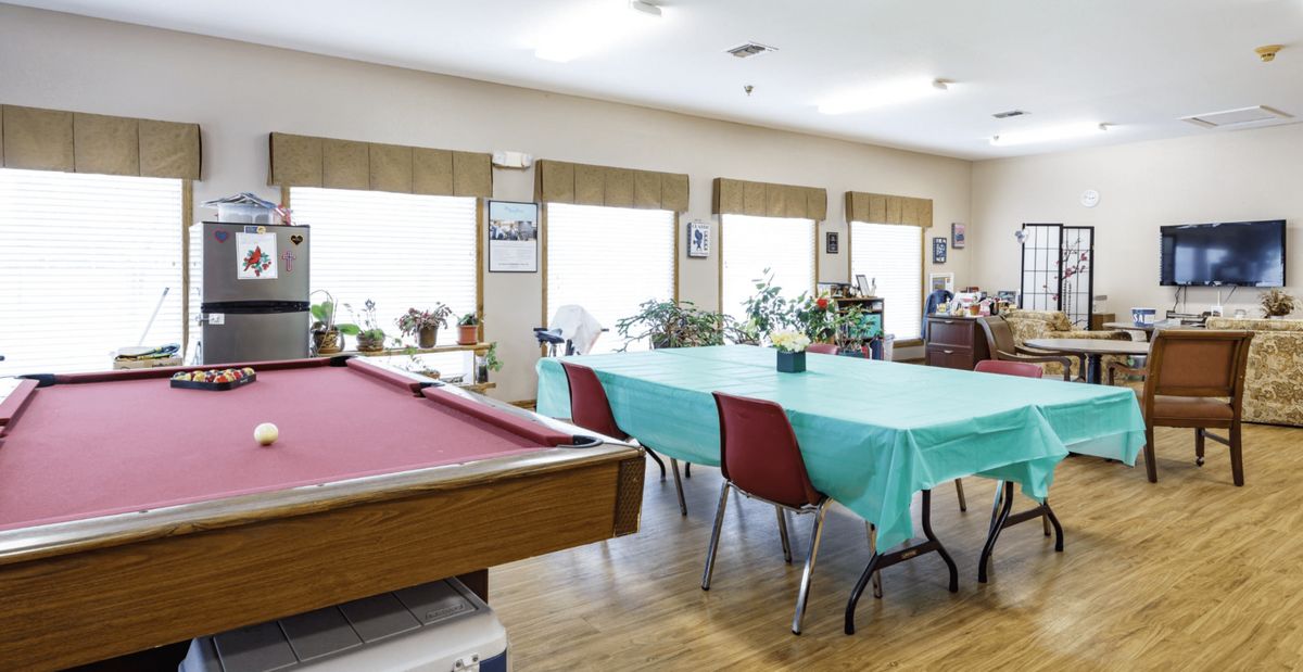 A recreation room featuring a billiards table and a dining area with tables and chairs, surrounded by windows and plants.
