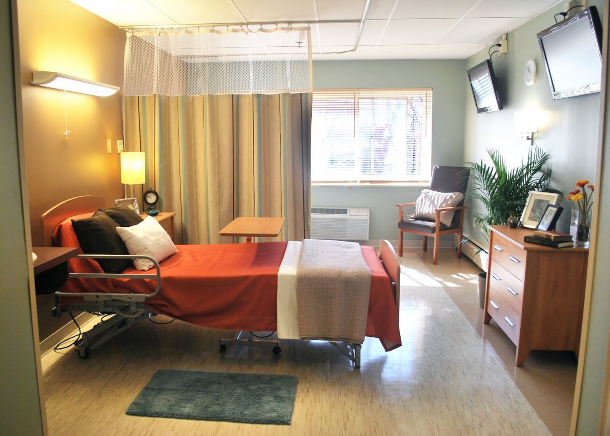 A modern hospital room with a single bed, a curtain for privacy, a window letting in natural light, and a wall-mounted television.