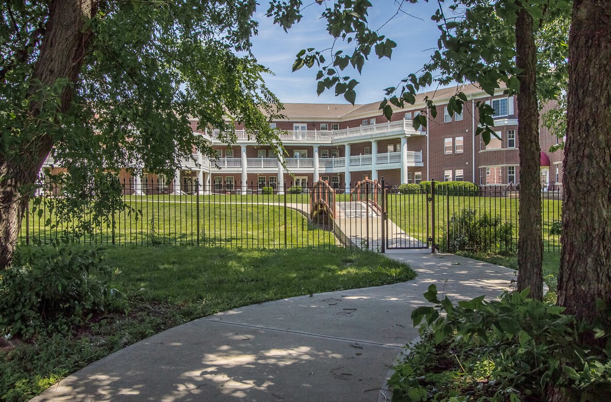 A pathway leads to a brick building with white columns, surrounded by greenery and enclosed by a fence.