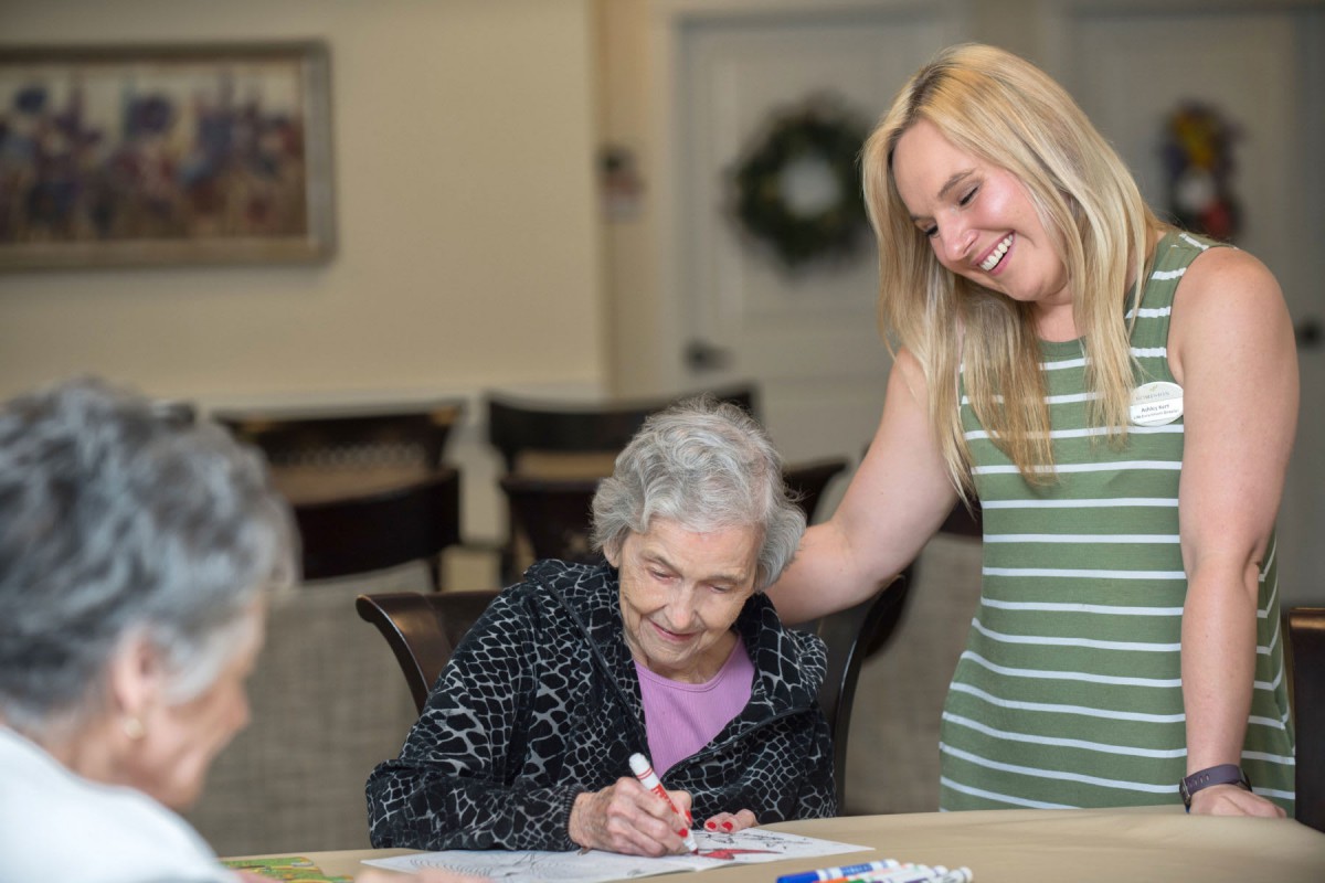 A young woman smiles as she interacts with an elderly woman engaged in an art activity.