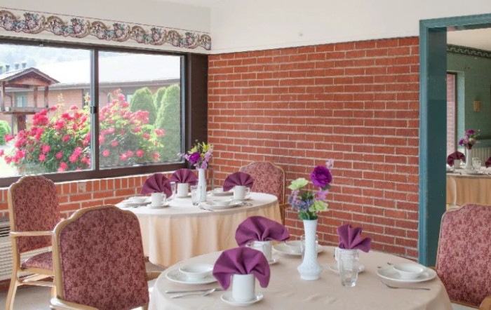 A cozy dining area with tables elegantly set with purple napkins and floral centerpieces, against a backdrop of a brick wall and window view of greenery.