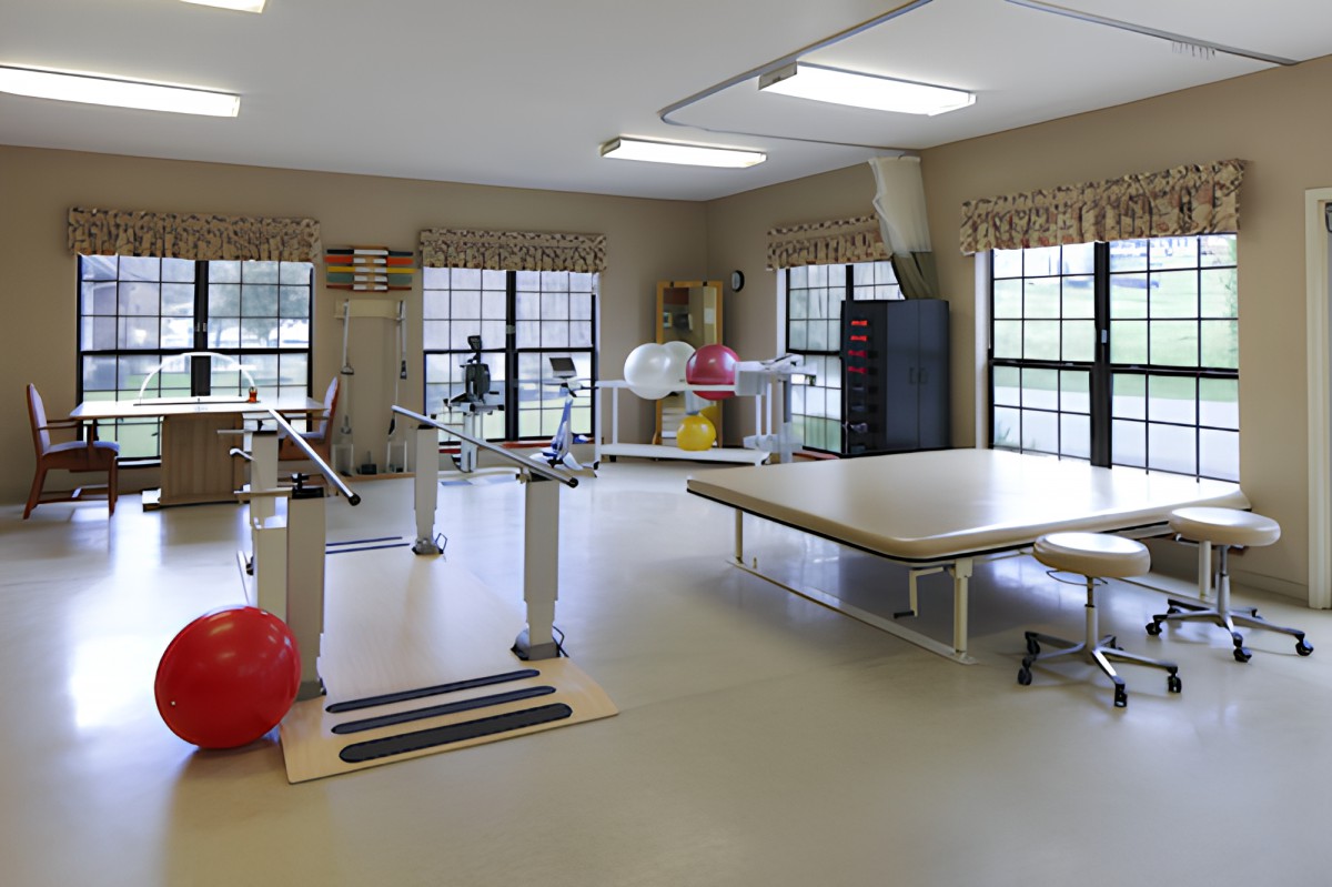 A well-equipped rehabilitation room with various physical therapy equipment, including a therapy table and gym balls, set in a bright space with large windows.