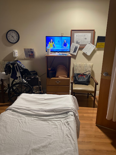 A view of a hospital room featuring a bed, TV, wheelchair, and various documents on the wall.
