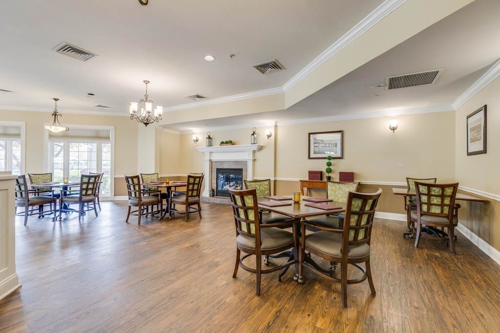 A spacious dining area with multiple tables, chairs, and a fireplace, featuring a wooden floor and bright lighting.