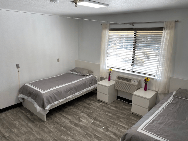 A minimalist bedroom with two single beds, grey bedding, and natural light from a window.