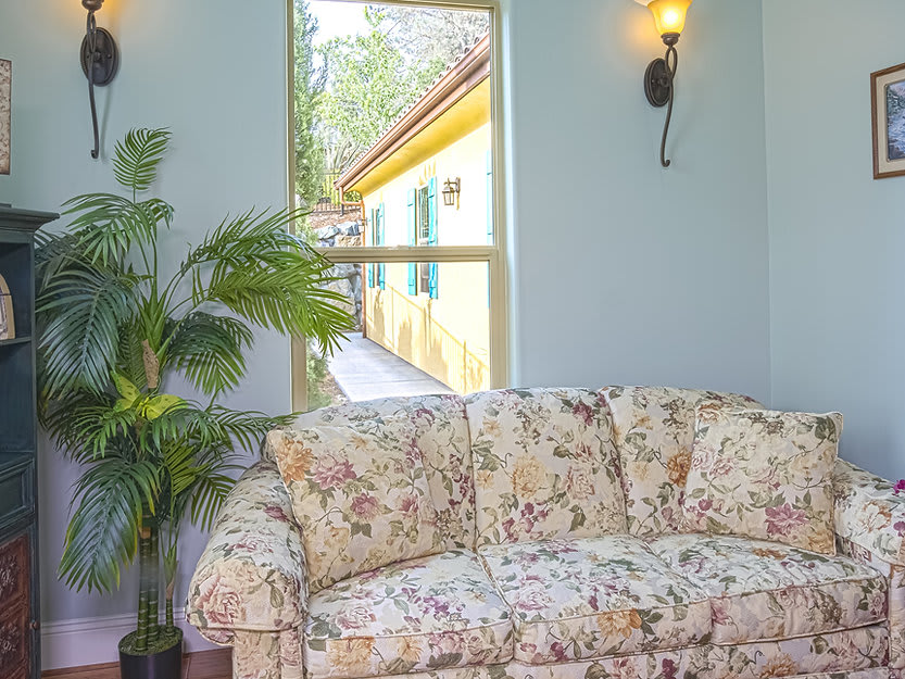 A cozy living room with a floral sofa, decorated with wall sconces and an indoor plant, offering a view through the window.