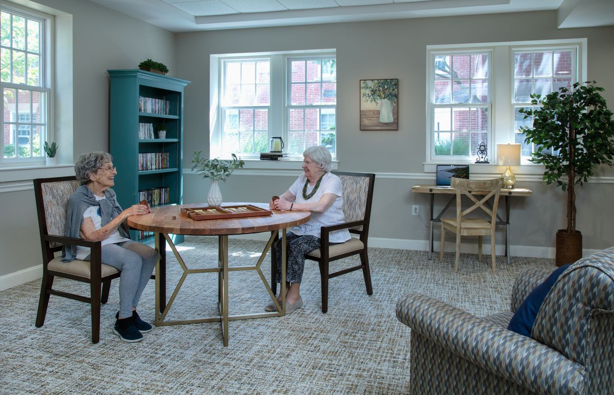 Two elderly individuals sitting in a cozy room, engaged in conversation at a table surrounded by bookshelves.